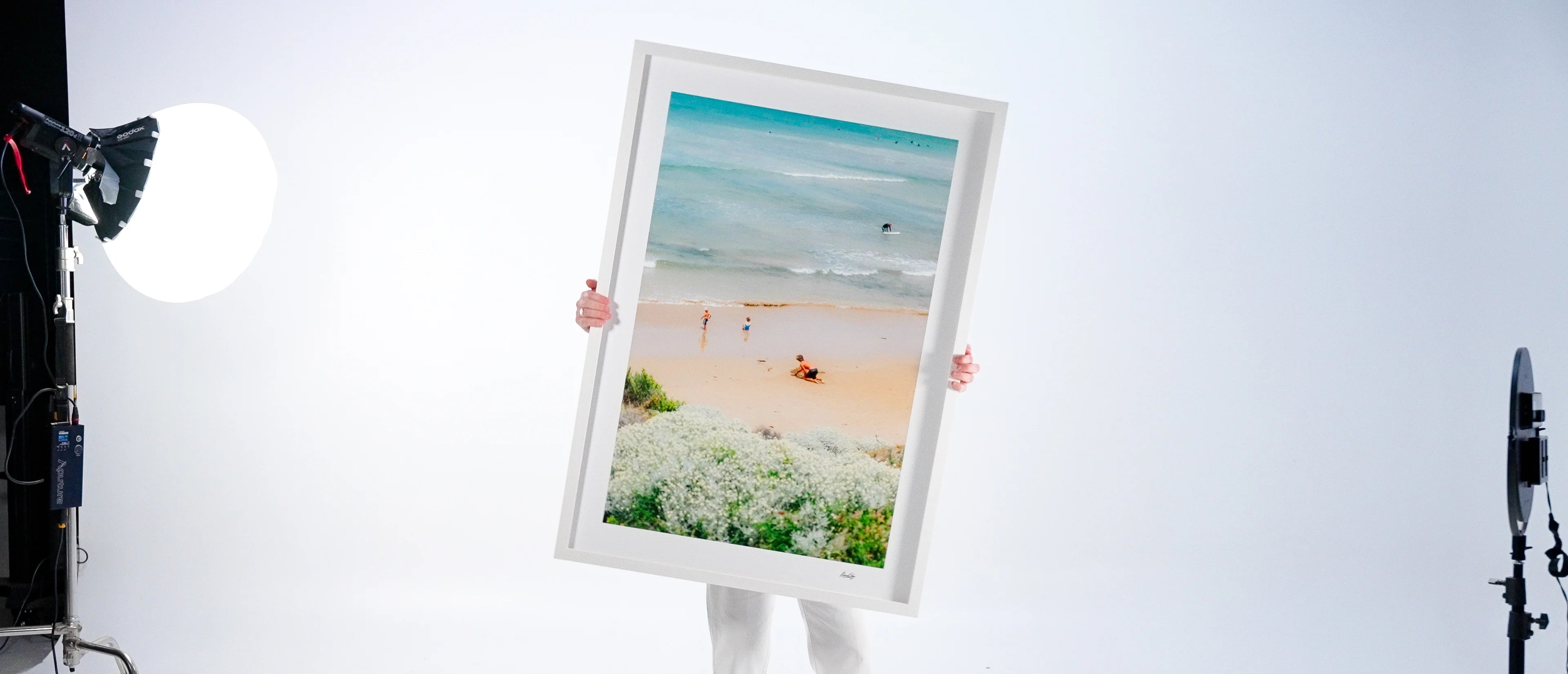Person holding a framed artwork of a beach scene in a studio setting with lighting equipment.